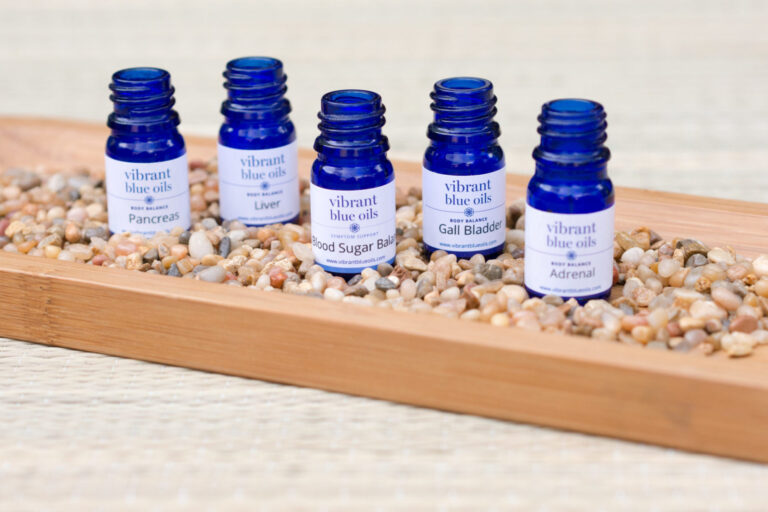 A selection of blue glass bottles with labels indicating various essential oils designed for different organs, such as pancreas, liver, blood sugar balance, gall bladder, and adrenal, displayed on a wooden tray nestled among small pebbles.