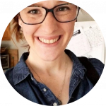 A person with glasses is smiling brightly in a selfie. They have light-colored hair tied back and are wearing a dark blue button-up shirt. A white refrigerator with notes and a bulletin board are visible in the background of a kitchen setting.
